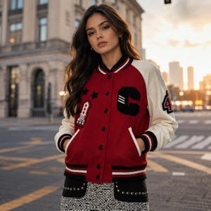 Può includere: Una giacca varsity rossa e crema con una lettera C nera sul petto, una toppa a forma di razzo con la scritta USA, tre stelle nere e il numero 44 sulla manica. La giacca presenta colletto, polsini e orlo a coste con strisce bianche e nere.