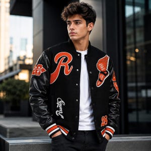 Può includere: Una giacca varsity nera con maniche in pelle nera, caratterizzata da patch in ciniglia arancione e bianco tra cui una grande lettera R, una sneaker alata, la scritta Runtz e il numero 28. La giacca presenta polsini a righe e una t-shirt bianca.