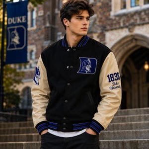 Może przedstawiać: Czarna kurtka typu varsity z beżowymi rękawami, ozdobiona niebieskim logo Duke University na piersi, liczbą 1838 oraz napisem Duke University na rękawie. Kurtka posiada zapięcie na zatrzaski oraz prążkowany kołnierz i mankiety z niebieskimi paskami.