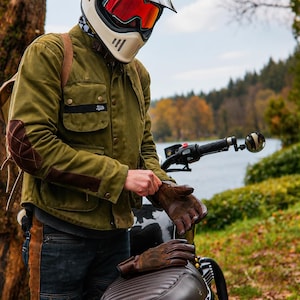 May include: An olive green waxed cotton motorcycle jacket with brown quilted elbow patches, worn with a cream-colored full-face helmet featuring a red visor, dark brown leather gloves, and dark denim jeans with brown leather side panels.