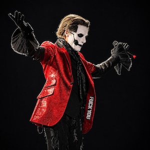 May include: A performer with white face paint and black eye markings wearing a red metallic blazer with the text FUCK YOU on the lapel, a black shirt, black scarf, and black gloves with ruffled cuffs, holding a microphone with arms outstretched.
