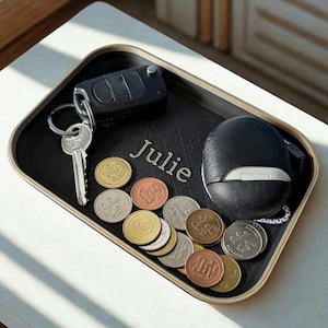 May include: A rectangular black valet tray with a tan border, featuring the name Julie engraved in the centre. The tray holds a black car key fob with a metal key, a black rounded earbud case, and a collection of various gold, silver, and copper coins.