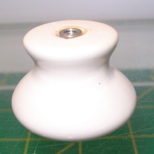May include: A white ceramic cabinet knob with a smooth, rounded, bulbous shape. The top of the knob features a flat surface with a centered, circular metal insert containing a threaded hole for mounting hardware, providing a functional design.