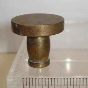 May include: A brass-toned metal knob with a flat circular top and a tapered cylindrical base. The knob is positioned next to a clear ruler, showing the top diameter is approximately 0.75 inches and the total height is approximately 1 inch.