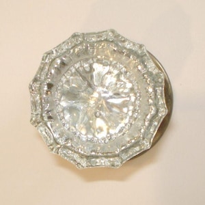 May include: A round, clear glass door knob featuring a faceted, multi-sided edge and a textured, starburst-like pattern radiating from the center. The knob is attached to a circular, metallic base plate with a tarnished, brass-toned finish.