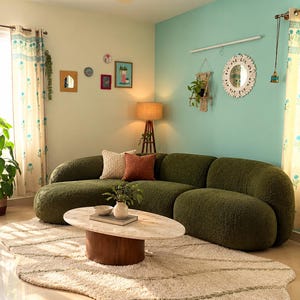 May include: A curved olive green textured sofa sits on a cream-colored rug with a wavy edge. In front is an oval marble coffee table with a wooden base, holding a small white vase with greenery and a bowl. Two throw pillows rest on the sofa.