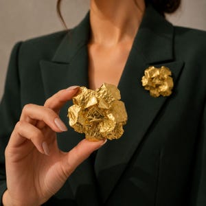 May include: A gold-toned brooch featuring a multi-layered, abstract floral design with a hammered, textured surface. The brooch has a rounded, sculptural shape with irregular, overlapping petals that create depth and dimension in a metallic finish.