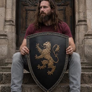 Op de afbeelding: Een schild in de vorm van een wapenschild met een matzwarte afwerking en een goudkleurig embleem van een klimmende leeuw in het midden. Het schild is omrand door een donkere metalen rand met gelijkmatig verdeelde decoratieve klinknagels.