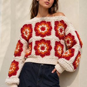 Könnte beinhalten: Ein langärmeliger Häkelpullover mit einer cremefarbenen Basis und einem sich wiederholenden Quadratmuster. Jedes Quadrat enthält ein mehrschichtiges Blumenmotiv in den Farben Dunkelrot, leuchtendes Orange und Weiß. Der Pullover hat einen weiten Ausschnitt und gerippte Bündchen.