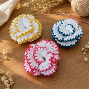 Può includere: Tre fiori lavorati all'uncinetto a mano con petali bianchi a strati. Un fiore presenta un bordo giallo, un altro un bordo blu scuro e il terzo un bordo rosa acceso. Ogni fiore è realizzato con un punto denso, strutturato e una forma rotonda.