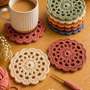 May include: A collection of round, crocheted coasters in various colors including sage green, cream, yellow, rust, and blue. A ceramic mug sits on a rust-colored coaster, while a pink 4.0 mm crochet hook and yarn balls are nearby. The text reads TheCrochetPrint.