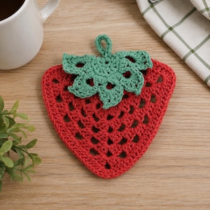 Puede incluir: Un posavasos o agarradera de ganchillo con forma de fresa, que presenta un cuerpo triangular rojo con un patrón de puntos calados y una parte superior de hojas verdes tejidas con un pequeño lazo para colgar. El artículo es de hilo texturizado.