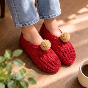 May include: A pair of deep red crochet slippers featuring a textured ribbed pattern and a single beige pom-pom attached to the top of each slipper. The slippers are worn with light blue denim jeans, showcasing a slip-on design with a rounded toe.