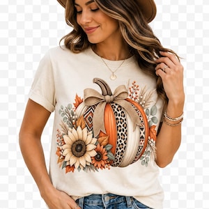 May include: A cream-colored short-sleeve t-shirt featuring a graphic of a pumpkin decorated with a bow, leopard print, and geometric patterns, surrounded by sunflowers and autumn foliage, paired with blue denim jeans and a brown felt fedora hat.