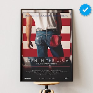 Puede incluir: Un póster enmarcado del álbum Born in the U.S.A. de Bruce Springsteen con una camiseta blanca y vaqueros azules sobre un fondo de rayas rojas y blancas. El texto dice Born in the U.S.A. Bruce Springsteen con una lista de canciones debajo.