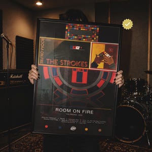 May include: A framed poster for The Strokes album Room on Fire, featuring a black background with red and blue geometric patterns, the band name, tracklist, and a square illustration of two hands holding a cigarette lighter in the upper right corner.