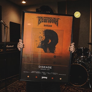 Puede incluir: Un póster rectangular enmarcado con la portada del álbum Disease de Beartooth. El póster tiene un fondo naranja con una silueta negra de un perfil de cabeza, el texto Beartooth Disease y una lista de canciones. El marco es negro y fino.