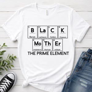 以下が含まれることがあります： 周期表の元素記号を組み合わせて「BLACK MOTHER」と綴ったグラフィックデザインがプリントされた白い半袖Tシャツ。元素記号の下には黒い大文字で「THE PRIME ELEMENT」と書かれています。クルーネックのTシャツです。