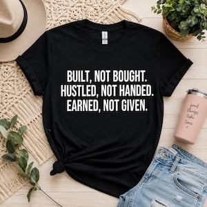 Könnte beinhalten: Ein schwarzes Kurzarm-T-Shirt mit weißem Text: BUILT, NOT BOUGHT. HUSTLED, NOT HANDED. EARNED, NOT GIVEN. Das T-Shirt hat einen Rundhalsausschnitt und liegt flach, wobei die fettgedruckte, zentrierte Typografie auf der Vorderseite deutlich zu sehen ist.