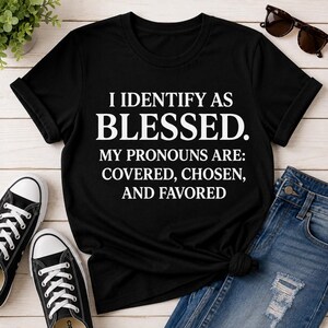 May include: A black short-sleeved t-shirt featuring white text that reads: I IDENTIFY AS BLESSED. MY PRONOUNS ARE: COVERED, CHOSEN, AND FAVORED. The shirt has a crew neckline and is displayed flat with a pair of blue jeans and black sunglasses.
