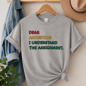 May include: A heather gray short-sleeve t-shirt featuring the text "DEAR ANCESTORS I UNDERSTAND THE ASSIGNMENT." The word "DEAR" is in red, "ANCESTORS" is in yellow, and "I UNDERSTAND THE ASSIGNMENT." is in green, all with black outlines.