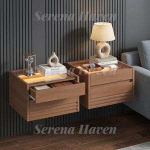 May include: Two wall-mounted wooden nightstands with glass tops and integrated warm lighting. Each unit features two drawers with horizontal slat detailing. The left nightstand has an open top drawer containing books and a glass hourglass.