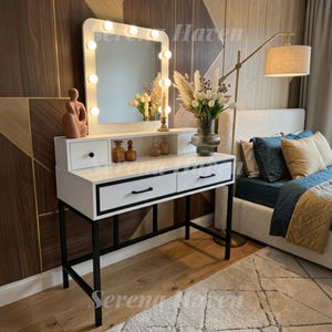 May include: A white vanity table with a black metal frame, featuring a rectangular mirror with integrated lights, two small upper drawers, and two large lower drawers with black handles. A terracotta sculpture and glass bottles sit on top.
