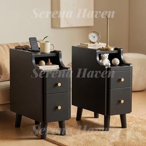 May include: Two black bedside tables with two drawers each, featuring gold-toned circular knobs and an open storage shelf at the top. The tables stand on four short legs and are positioned side by side, displaying a sleek, vertical design.