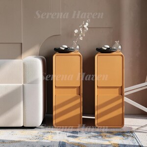 May include: Two tall, rectangular, mustard-yellow storage cabinets with rounded corners, each featuring two stacked square cabinet doors. On top of each cabinet sits a black circular tray containing white stones and a clear glass vase.