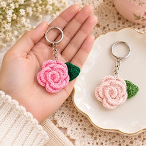 May include: Two crochet rose keychains featuring pink yarn flowers with small green leaves attached. Each flower is connected to a silver-toned metal split ring by a short chain. One keychain is held in a palm, while the other rests nearby.