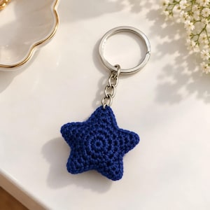 Può includere: Un portachiavi a forma di stella blu scuro realizzato in filo lavorato all'uncinetto. La stella è fissata a un anello metallico color argento tramite una corta catena di maglie metalliche. La trama mostra il motivo complesso dei punti.