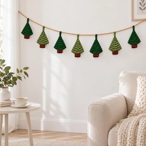 May include: A decorative garland featuring eight crocheted Christmas tree ornaments in varying shades of green, each with a brown trunk, suspended from a single tan twine string against a plain white wall.