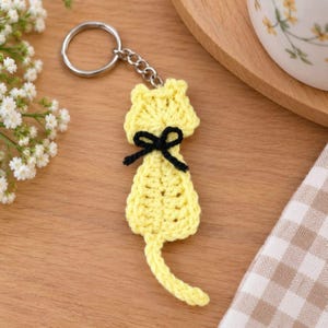 May include: A yellow crocheted cat keychain featuring a small black yarn bow tied around the neck area. The cat figure is attached to a silver-toned metal split ring by a short chain, showcasing a textured yarn stitch pattern throughout.