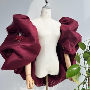 May include: A burgundy bolero jacket featuring voluminous, oversized puff sleeves made of sheer fabric. The garment is displayed on a mannequin and includes intricate dark red floral embellishments with small black beads on the sleeves.