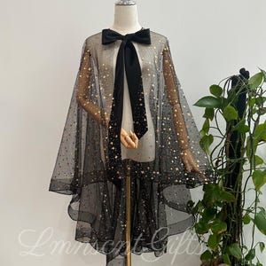 Peut inclure: Une cape noire transparente ornée d'un motif de petites étoiles dorées, présentée sur un mannequin. La cape possède un col haut avec un grand nœud en tissu noir uni noué au centre. Le tissu est léger, translucide et superposé.