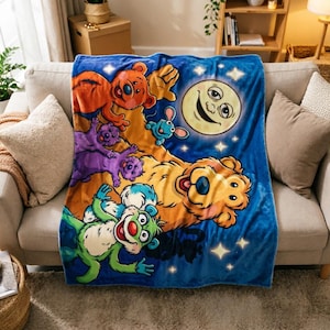 May include: A soft blue fleece blanket featuring a large yellow bear, a smaller orange bear, a green creature, and a purple creature. The design includes a smiling moon with a face and several white stars against a dark blue night sky background.
