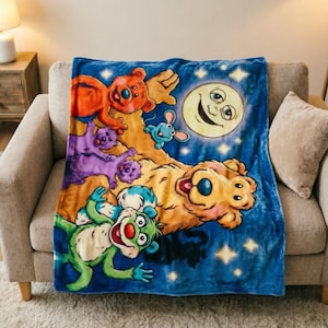 Pode incluir: Uma manta de lã azul macia com um grande urso amarelo sorridente, um urso verde, um urso laranja e dois pequenos ursos roxos. O design inclui uma lua sorridente e estrelas amarelas sobre um fundo de céu noturno azul escuro.