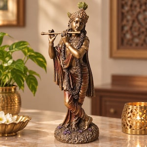 May include: A bronze-toned statue of a standing figure playing a flute. The figure wears an ornate crown with a peacock feather, layered necklaces, and draped garments. The statue is positioned on a textured base with small purple floral accents.