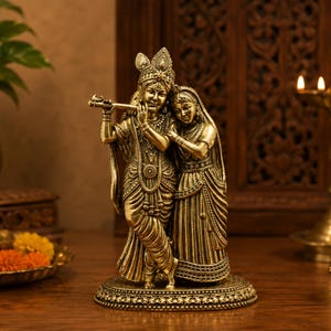 May include: A metallic gold-toned statue depicting two figures standing together on an oval base. One figure holds a flute to their lips while the other leans against them. Both figures wear traditional ornate clothing, jewellery, and headpieces.