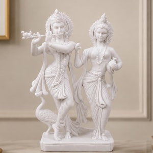 May include: A white marble-finish statue depicting two figures standing together on a rectangular base. One figure plays a flute while the other stands beside them. Both figures wear ornate headpieces, necklaces, and draped garments. A peacock stands at the base.