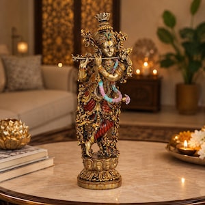 Puede incluir: Una estatua detallada en tono dorado de la deidad Krishna tocando una flauta. La figura lleva una corona ornamentada, joyas y una prenda roja con detalles florales azules y rosas. La estatua se asienta sobre una base circular en forma de loto.