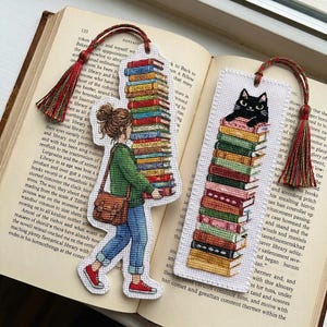 以下が含まれることがあります： タッセル付きの刺繍しおり2点。1点は緑のセーターと青いジーンズを着た人物が本を高く積み上げて運んでいる様子。もう1点は長方形で、カラフルな本の山の上に座る黒猫が刺繍されている。