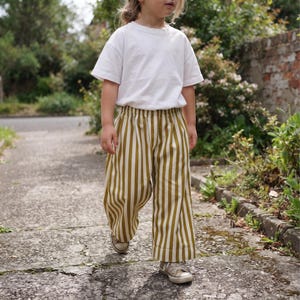 May include: A plain white short-sleeved t-shirt paired with loose-fitting trousers featuring vertical mustard yellow and white stripes. The trousers have an elasticated waistband and a wide-leg cut, worn with low-top canvas sneakers.