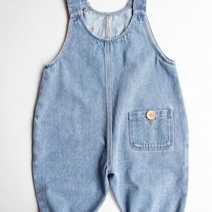 Könnte beinhalten: Eine hellblaue Latzhose aus Denim mit rundem Ausschnitt, breiten Trägern und einer aufgesetzten Tasche auf der Vorderseite. Die Tasche ist mit einem runden, hellen Holzknopf verziert. Die Hosenbeine sind mit umgeschlagenen Säumen versehen.