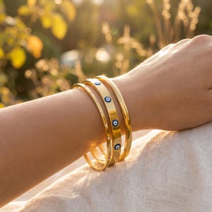 May include: A set of three gold-toned metal bangles. One bangle features a series of small, circular blue and white evil eye motifs. Another bangle is adorned with a row of tiny, clear embedded stones. The third bangle is a plain, smooth gold band.