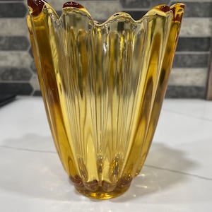 Op de afbeelding: Een doorschijnende amberkleurige glazen vaas met een verticaal geribbeld patroon en een geschulpte, golvende rand. De vaas heeft een brede, uitlopende opening die taps toeloopt naar een kleinere, ronde voet, met een consistente goudgele kleur.