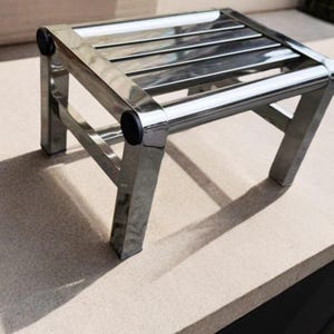 May include: A small, rectangular step stool constructed from polished, reflective stainless steel. The stool features a slatted top surface and four sturdy legs, each accented with a circular black plastic cap at the top corner joints.