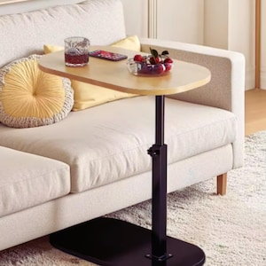 Op de afbeelding: Een in hoogte verstelbare bijzettafel met een ovaal tafelblad in een lichte houtkleur en een zwart metalen onderstel. De tafel heeft een centrale verticale steunkolom met een handmatige instelknop op een platte, ovale zwarte voet.