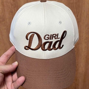 May include: A baseball cap featuring a cream-colored crown and a brown brim. The front of the cap displays the embroidered text "Girl Dad" in a brown, cursive script font. The cap includes a small brown button at the top and stitched eyelets.
