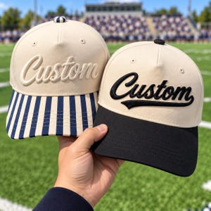 May include: Two baseball caps with cream-colored crowns and the word Custom embroidered on the front. One cap features a navy blue and white vertical striped brim with a striped button, while the other has a solid black brim and black button.